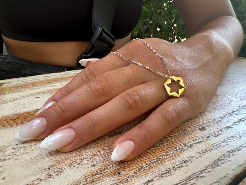 May include: A gold necklace with a hexagonal pendant featuring a star of David cutout. The necklace is worn by a person with white-tipped nails.