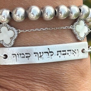 Op de afbeelding: Zilveren armbanden met verschillende ontwerpen. E&eacute;n armband heeft grote, ronde kralen. Een andere heeft een ketting met een rechthoekige plaat met gegraveerde tekst. Een derde armband heeft een klaverontwerp met een wit midden.