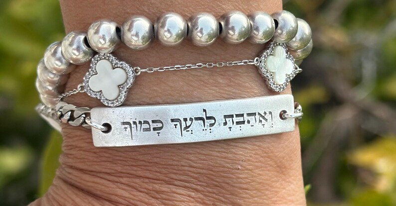 Op de afbeelding: Zilveren armbanden met verschillende ontwerpen. E&eacute;n armband heeft grote, ronde kralen. Een andere heeft een ketting met een rechthoekige plaat met gegraveerde tekst. Een derde armband heeft een klaverontwerp met een wit midden.