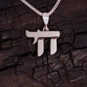 May include: A silver Chai pendant necklace. The pendant features the Hebrew letter Chai, meaning "life", in a stylized design. The necklace has a delicate chain and is displayed against a dark, textured background.