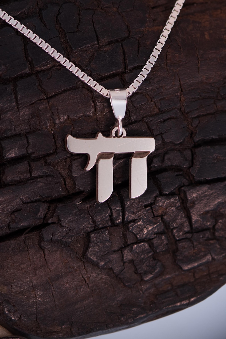 May include: A silver Chai pendant necklace. The pendant features the Hebrew letter Chai, meaning "life", in a stylized design. The necklace has a delicate chain and is displayed against a dark, textured background.