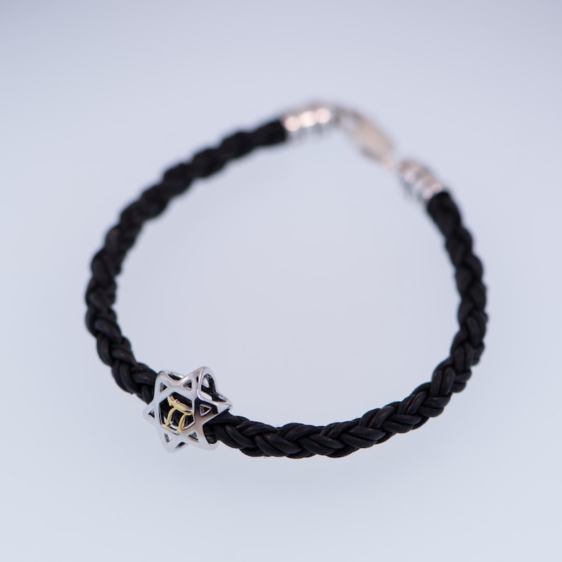 K&ouml;nnte beinhalten: Ein schwarzes geflochtenes Lederarmband mit einem silbernen Davidstern-Anh&auml;nger mit einer goldenen hebr&auml;ischen Inschrift.