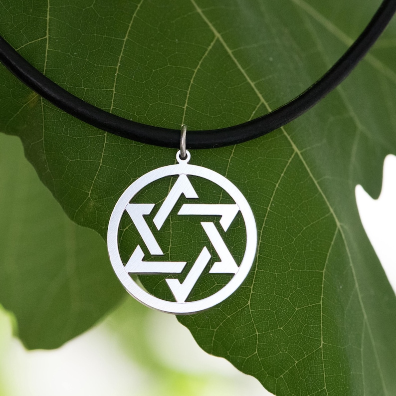 Pu&ograve; includere: Una collana con pendente in argento con un design a Stella di David. Il pendente &egrave; circolare con una stella a sei punte ritagliata al centro. La collana ha un cordino nero. Lo sfondo &egrave; una foglia verde.