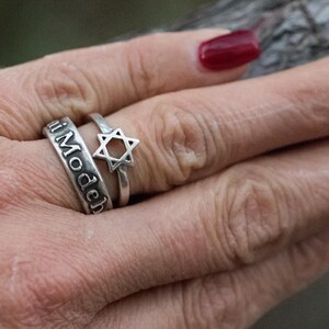 Puede incluir: Se muestran dos anillos de plata en una mano. Un anillo tiene grabadas las palabras "i Modech". El otro anillo presenta un dise&ntilde;o de Estrella de David. La mano tiene esmalte de u&ntilde;as rojo.