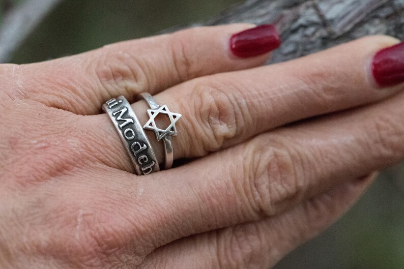 Puede incluir: Se muestran dos anillos de plata en una mano. Un anillo tiene grabadas las palabras "i Modech". El otro anillo presenta un dise&ntilde;o de Estrella de David. La mano tiene esmalte de u&ntilde;as rojo.