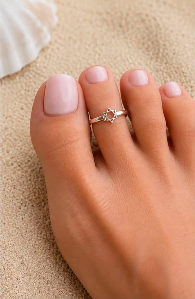 Puede incluir: Un anillo para el dedo del pie de tono plateado que presenta un peque&ntilde;o dije de la Estrella de David con marco abierto centrado en una banda de metal fino y pulido. El anillo se lleva en el segundo dedo, con esmalte rosa claro.