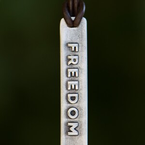 May include: A silver rectangular pendant with the word "FREEDOM" embossed vertically. The pendant is suspended from a brown leather cord. The background is a blurred, dark green.
