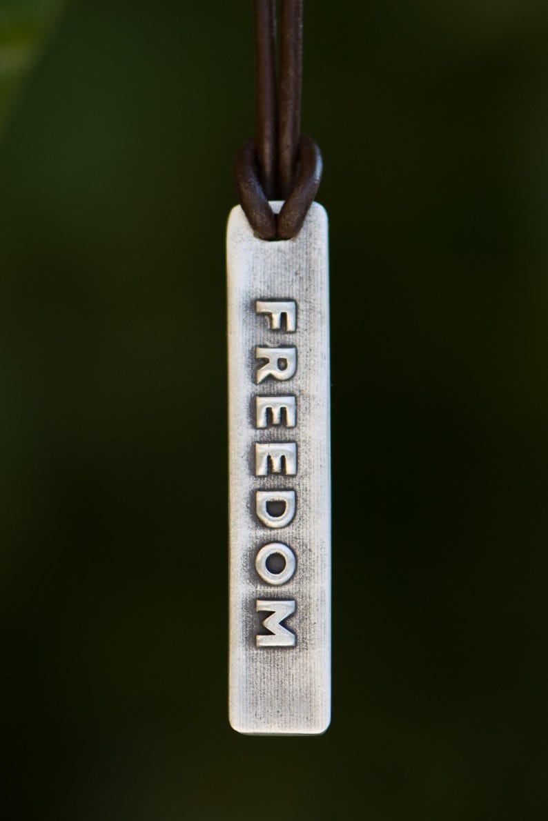 May include: A silver rectangular pendant with the word "FREEDOM" embossed vertically. The pendant is suspended from a brown leather cord. The background is a blurred, dark green.