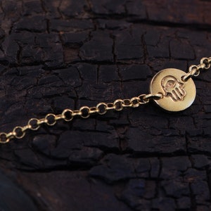 May include: A gold chain bracelet with a round charm featuring a hamsa hand design. The charm has a small eye in the center of the palm.