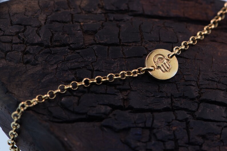 May include: A gold chain bracelet with a round charm featuring a hamsa hand design. The charm has a small eye in the center of the palm.