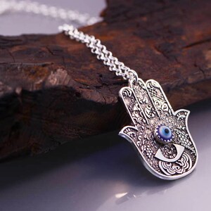 May include: Silver necklace with a hamsa hand charm featuring a blue evil eye. The hamsa hand is a symbol of protection and good luck.