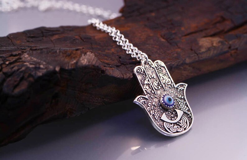 May include: Silver necklace with a hamsa hand charm featuring a blue evil eye. The hamsa hand is a symbol of protection and good luck.