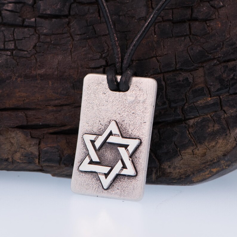 May include: Silver-tone rectangular pendant necklace featuring a Star of David design. The pendant hangs from a black leather cord. The Star of David is a six-pointed star, a symbol of Judaism.
