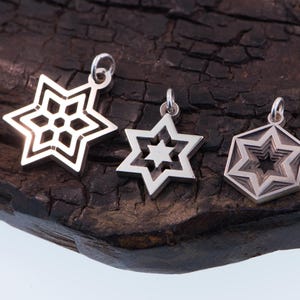 May include: Three silver star pendants with varying designs. One features an openwork snowflake, another a six-pointed star, and the third a layered star within a hexagon. They are displayed on a dark, textured wooden surface.