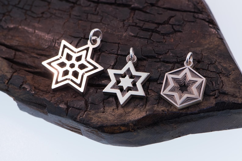May include: Three silver star pendants with varying designs. One features an openwork snowflake, another a six-pointed star, and the third a layered star within a hexagon. They are displayed on a dark, textured wooden surface.