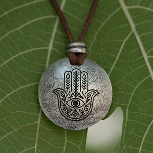 May include: A silver-colored pendant necklace featuring a Hamsa hand design with an eye in the center. The pendant hangs from a brown cord. The necklace is displayed on a green leaf.