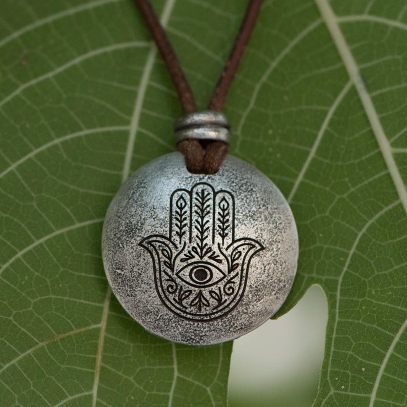 May include: A silver-colored pendant necklace featuring a Hamsa hand design with an eye in the center. The pendant hangs from a brown cord. The necklace is displayed on a green leaf.