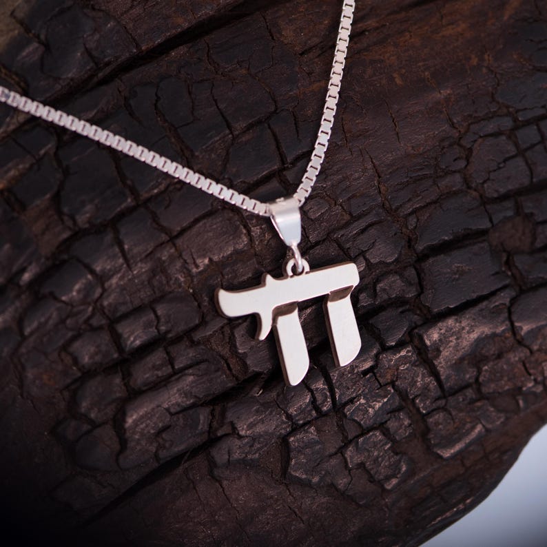 May include: A silver pendant necklace with the Hebrew letter "Chai". The pendant is suspended from a fine chain. The necklace is set against a dark, textured wooden backdrop.