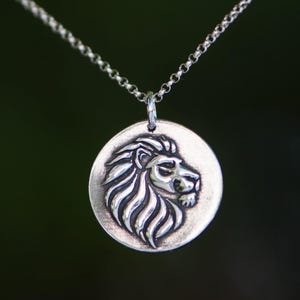 May include: A silver pendant necklace featuring a detailed lion's head in profile. The lion's mane is intricately carved, and the pendant hangs from a delicate chain. The background is a blurred, dark green.