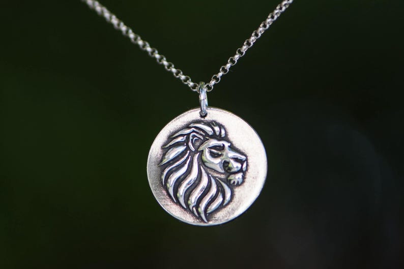 May include: A silver pendant necklace featuring a detailed lion's head in profile. The lion's mane is intricately carved, and the pendant hangs from a delicate chain. The background is a blurred, dark green.