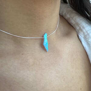 Op de afbeelding: Een zilveren ketting met een blauwe opaal hanger. De opaal is druppelvormig en heeft een iriserende glans.