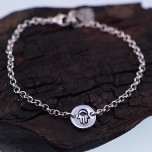 May include: A silver chain bracelet with a hamsa hand charm. The charm is round and has a raised hamsa hand design with an eye in the center.