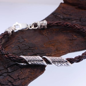 Pu&ograve; includere: Un braccialetto intrecciato in pelle marrone con un ciondolo attorcigliato in argento. Il ciondolo &egrave; inciso con dettagli intricati.