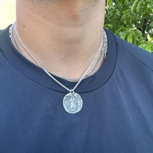 Pode incluir: Um colar prateado com um pingente de le&atilde;o. O pingente redondo apresenta um rosto de le&atilde;o detalhado. O colar &eacute; composto por duas correntes diferentes. A pessoa est&aacute; vestindo uma camisa azul escura.