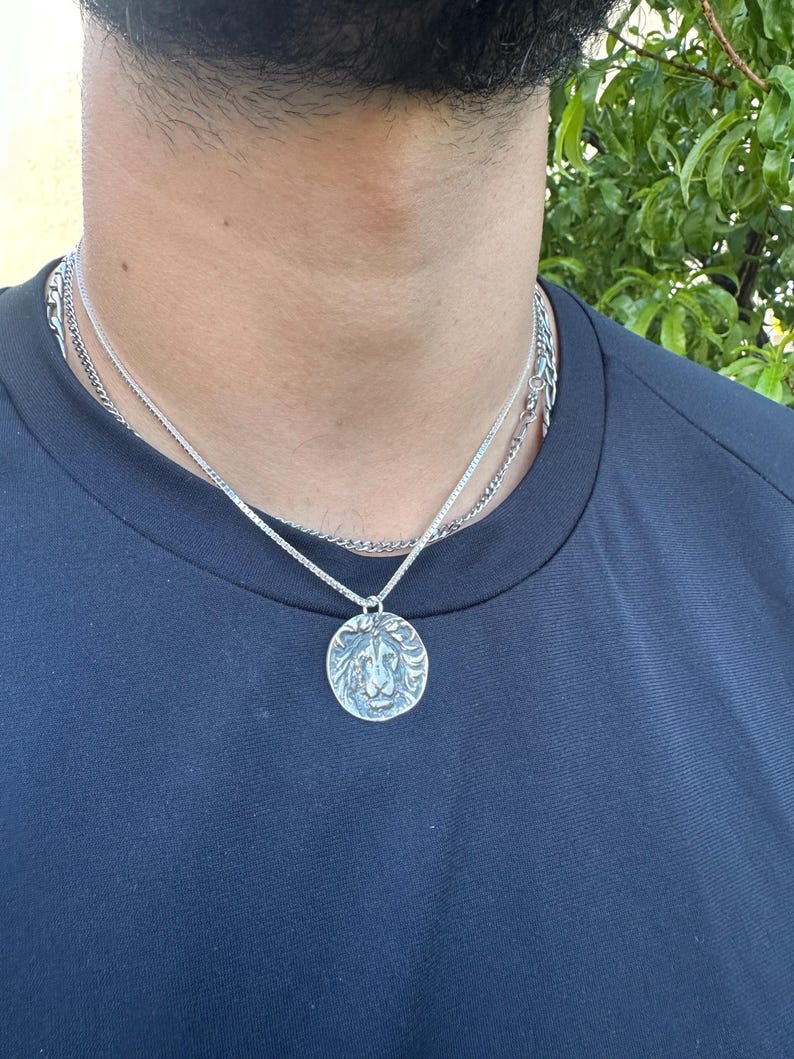 Pode incluir: Um colar prateado com um pingente de le&atilde;o. O pingente redondo apresenta um rosto de le&atilde;o detalhado. O colar &eacute; composto por duas correntes diferentes. A pessoa est&aacute; vestindo uma camisa azul escura.