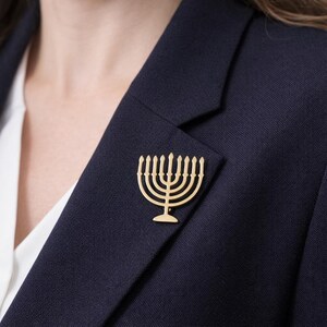 K&ouml;nnte beinhalten: Eine goldfarbene Menora-Brosche ist an einem marineblauen Blazer befestigt. Die Brosche ist eine stilisierte Darstellung des j&uuml;dischen Leuchters mit neun Kerzenhaltern. Der Blazer wird &uuml;ber einer wei&szlig;en Bluse getragen.