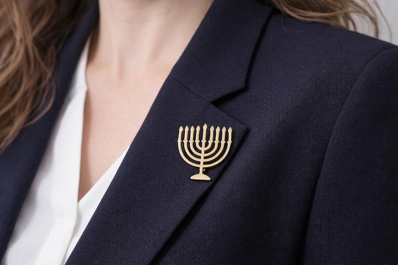 K&ouml;nnte beinhalten: Eine goldfarbene Menora-Brosche ist an einem marineblauen Blazer befestigt. Die Brosche ist eine stilisierte Darstellung des j&uuml;dischen Leuchters mit neun Kerzenhaltern. Der Blazer wird &uuml;ber einer wei&szlig;en Bluse getragen.