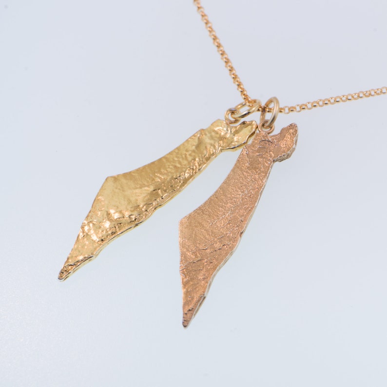 May include: A gold-toned necklace featuring two distinct map-shaped pendants suspended from a delicate chain. One pendant has a bright, polished gold finish with a textured surface, while the other has a matte, rose-gold appearance with a rougher texture.