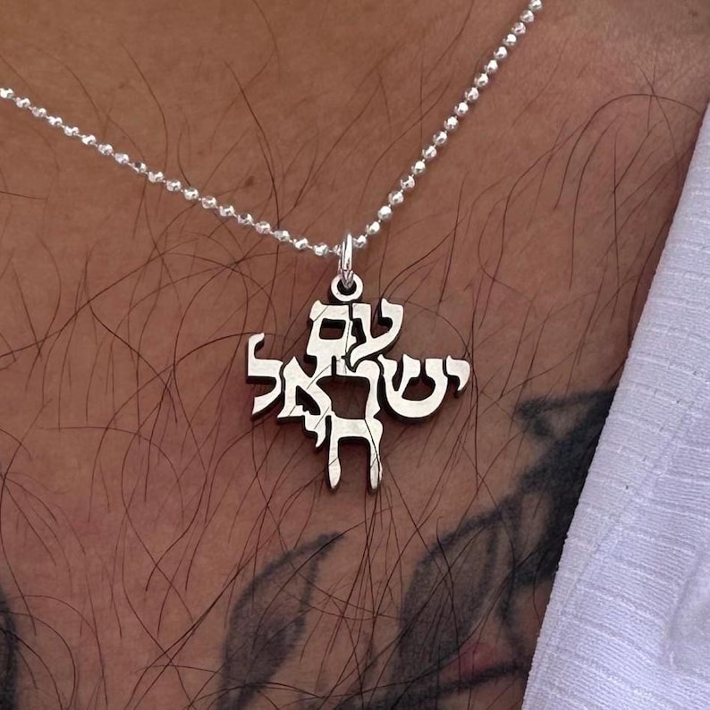 Puede incluir: Un collar de plata con un colgante con letras hebreas. El colgante tiene forma de cruz.