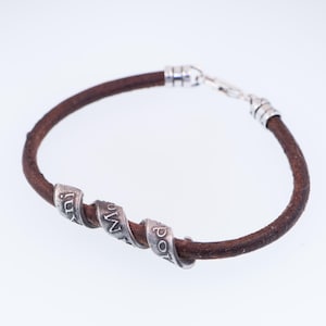 May include: A brown leather bracelet with three silver beads, each engraved with a word. The bracelet has a silver clasp. The leather band is approximately 0.6 cm in diameter.