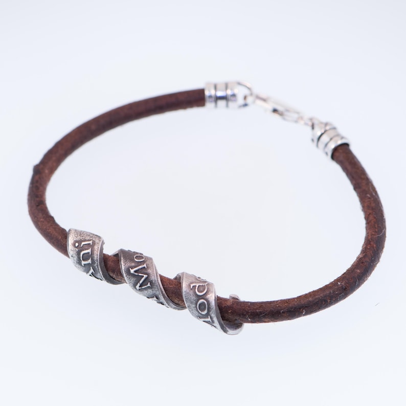 May include: A brown leather bracelet with three silver beads, each engraved with a word. The bracelet has a silver clasp. The leather band is approximately 0.6 cm in diameter.