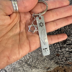 K&ouml;nnte beinhalten: Ein silberner Schl&uuml;sselanh&auml;nger mit einem Davidstern-Anh&auml;nger und einem rechteckigen Anh&auml;nger. Der Anh&auml;nger ist mit einem Davidstern, einer Karte Israels und dem hebr&auml;ischen Wort "Chai" graviert. Der Schl&uuml;sselanh&auml;nger wird in einer Hand gehalten.