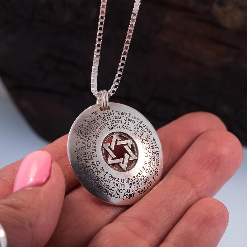 Puede incluir: Collar con colgante de plata con un colgante circular con texto grabado y un dise&ntilde;o de Estrella de David. El colgante tiene un centro rojo y cuelga de una delicada cadena de plata. El collar se sostiene en una mano.