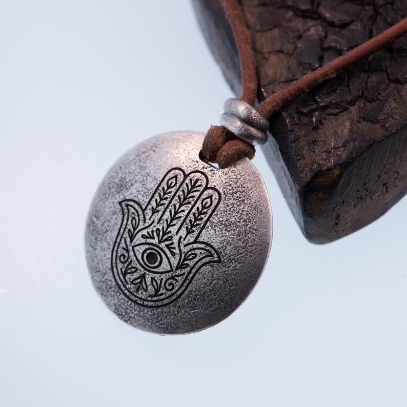 May include: A silver-colored pendant necklace featuring a Hamsa hand design with an eye in the center. The pendant hangs from a brown leather cord. The necklace has a rustic, textured appearance.