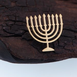 K&ouml;nnte beinhalten: Eine goldfarbene Menora, ein j&uuml;discher Leuchter, wird auf einer dunklen, strukturierten Holzoberfl&auml;che gezeigt. Die Menora hat neun Kerzenhalter und einen zentralen Stiel, der die acht Tage von Chanukka plus die Schamasch-Kerze darstellt.