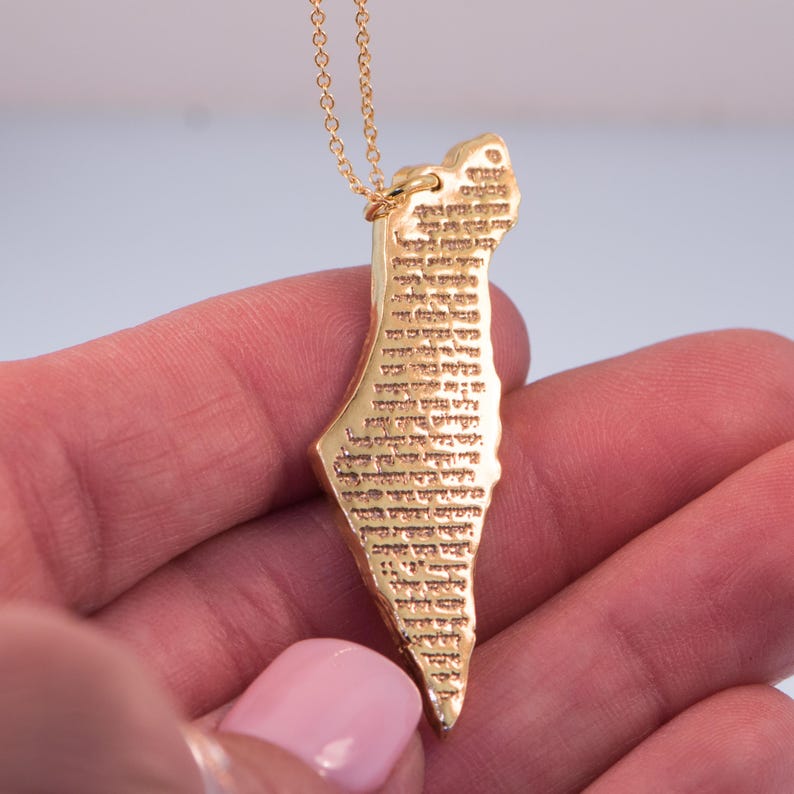 May include: A gold-colored pendant necklace in the shape of a map of Israel, with detailed text etched on the surface. The pendant is suspended from a delicate gold chain. The necklace is held in a person's hand.