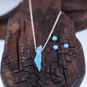 Op de afbeelding: Een zilveren ketting met een blauwe opaal hanger in de vorm van de omtrek van Isra&euml;l. Drie kleinere blauwe opaal stenen zijn verspreid over een donker houten oppervlak.
