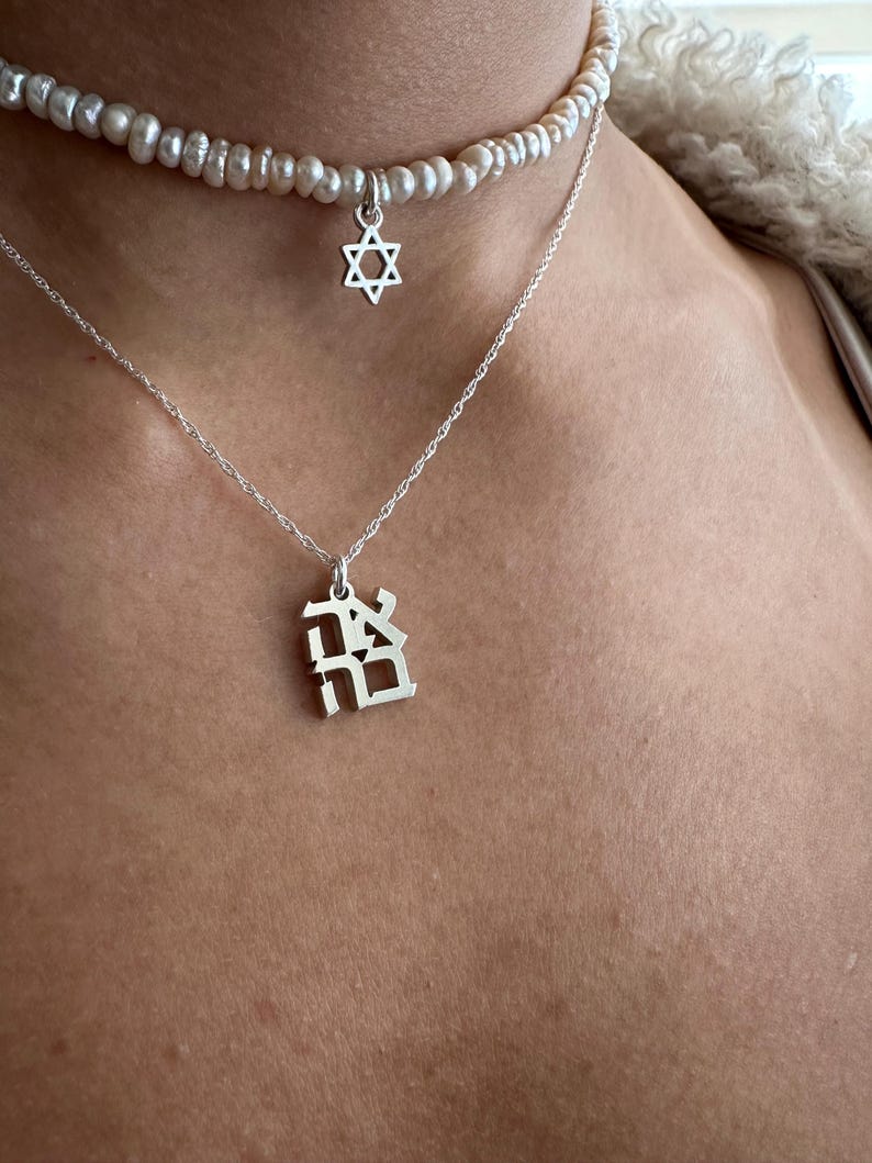 May include: Two silver necklaces with pendants. The top necklace is a pearl choker with a silver Star of David pendant. The bottom necklace has a silver pendant with a Hebrew letter.