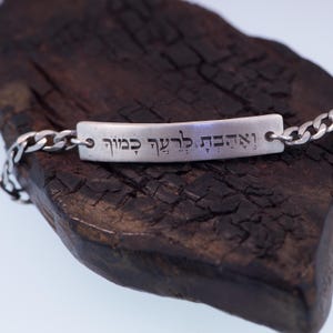 Op de afbeelding: Een zilveren armband met een rechthoekige balk gegraveerd met Hebreeuwse tekst. De armband heeft een schakelketting ontwerp en wordt gepresenteerd op een donker, getextureerd houten oppervlak. De Hebreeuwse tekst is duidelijk zichtbaar.