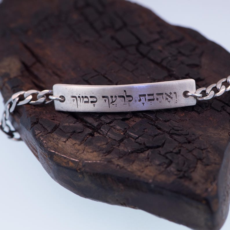 Op de afbeelding: Een zilveren armband met een rechthoekige balk gegraveerd met Hebreeuwse tekst. De armband heeft een schakelketting ontwerp en wordt gepresenteerd op een donker, getextureerd houten oppervlak. De Hebreeuwse tekst is duidelijk zichtbaar.