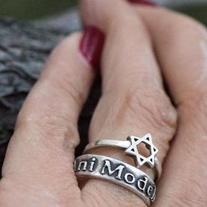 Puede incluir: Dos anillos de plata apilados en un dedo. El anillo superior presenta una Estrella de David, mientras que el anillo inferior tiene la inscripci&oacute;n "ni Mode". Los anillos se colocan sobre un fondo borroso.