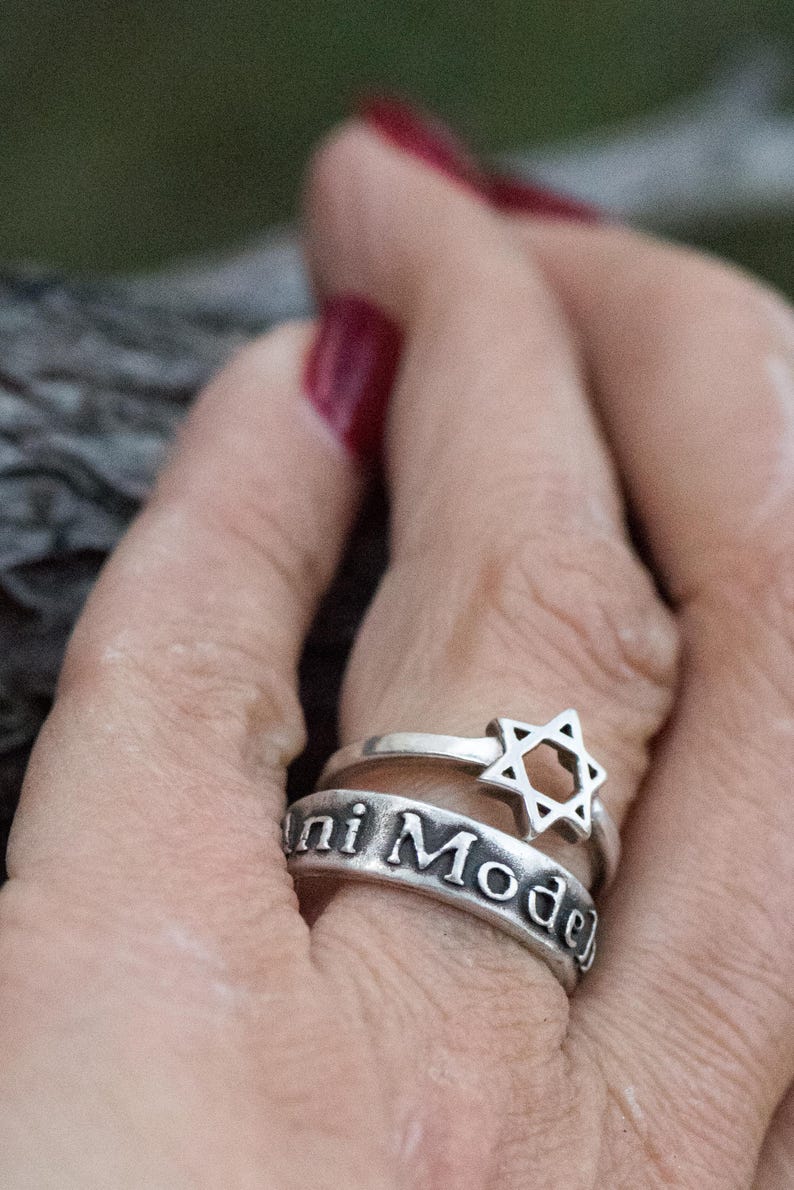 Puede incluir: Dos anillos de plata apilados en un dedo. El anillo superior presenta una Estrella de David, mientras que el anillo inferior tiene la inscripci&oacute;n "ni Mode". Los anillos se colocan sobre un fondo borroso.