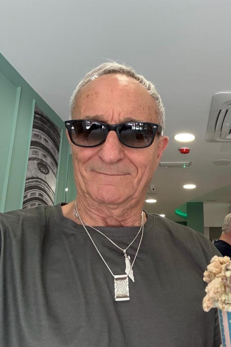 May include: A person wearing black sunglasses and a dark grey t-shirt. Two silver necklaces are visible, one featuring a rectangular pendant with engraved text and the other a smaller, pointed silver charm. The person has short, light-coloured hair.