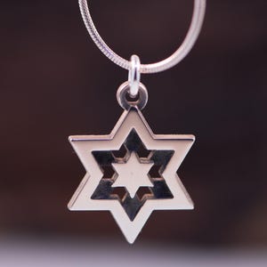 May include: A silver Star of David pendant necklace. The pendant has a six-pointed star design with a smaller star in the centre. The necklace chain is a thin, silver snake chain. The background is a blurred, dark brown.