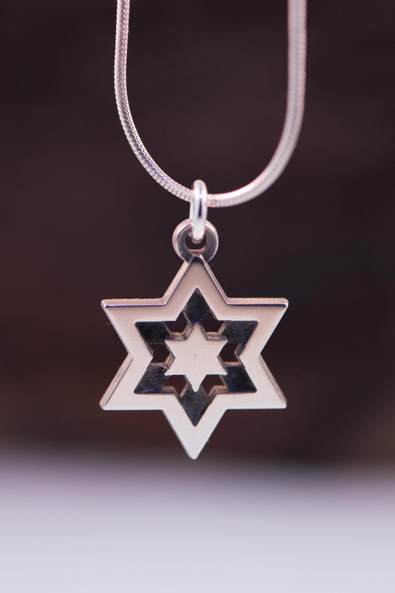 May include: A silver Star of David pendant necklace. The pendant has a six-pointed star design with a smaller star in the centre. The necklace chain is a thin, silver snake chain. The background is a blurred, dark brown.