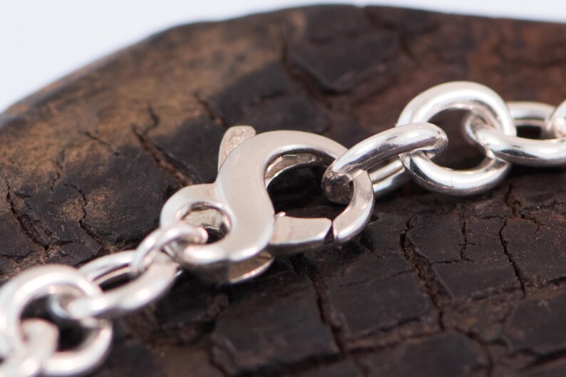 May include: A silver chain necklace featuring a polished lobster claw clasp. The metal links are round and interconnected, creating a continuous loop. The clasp mechanism is visible in the centre, showing the spring-loaded lever and the hook.